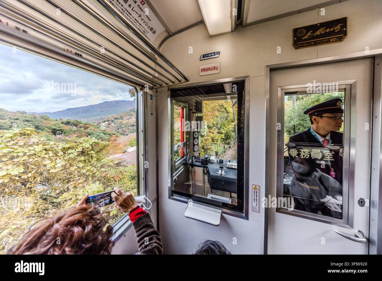 A passenger photographs the autumn mountain scenery from the window of the Hakone Tozan Railway (箱根登山鉄道) with her phone. A conductor sits in his cabin in the background. A plaque in the carriage bears the name St. Moritz. Since 1979 a partnership has linked the Bernina line of Switzerland's Rhaetian Railway with the Hakone Tozan Railway. In 2014 Hakone and St. Moritz also became sister cities. Hakone, Kanagawa Prefecture, Japan. Stock Photo