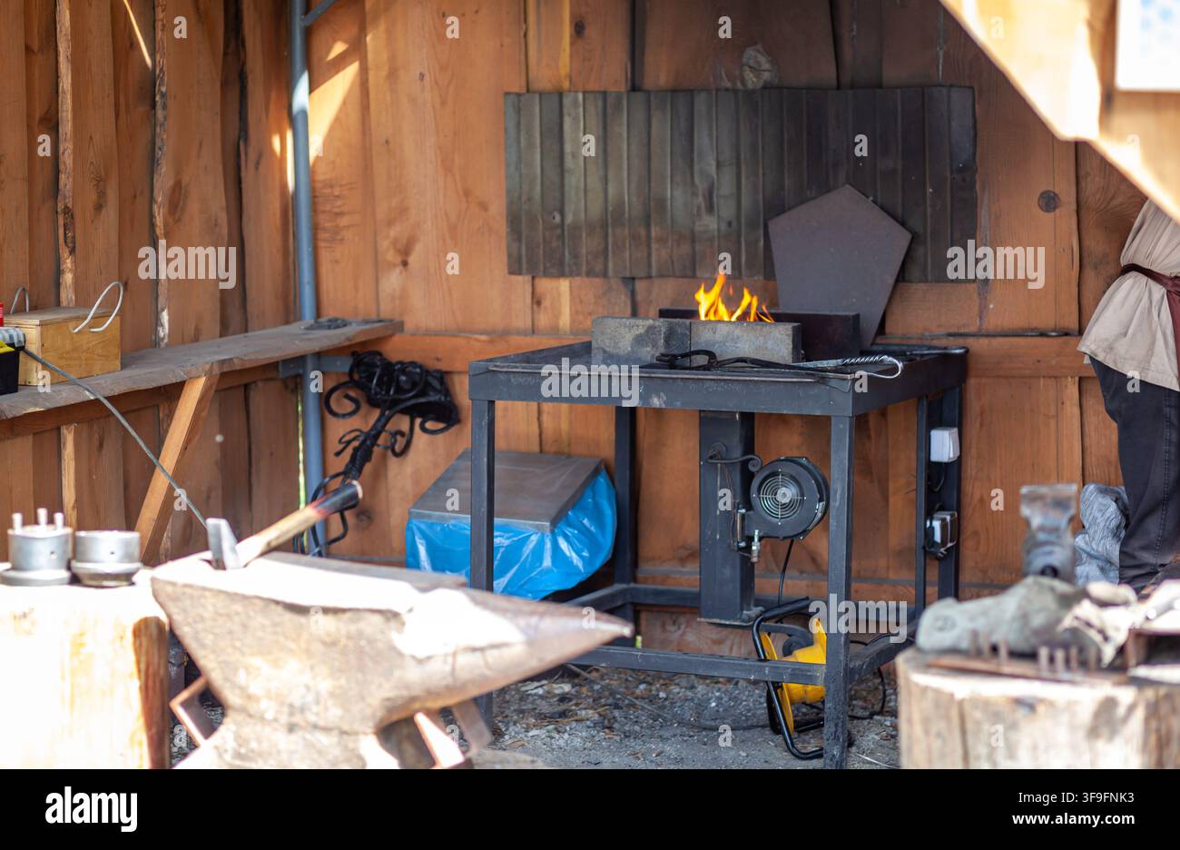 Blacksmith workshop interior featuring an anvil, forge with flames, and various tools on a workbench in a rustic wooden setting Stock Photo