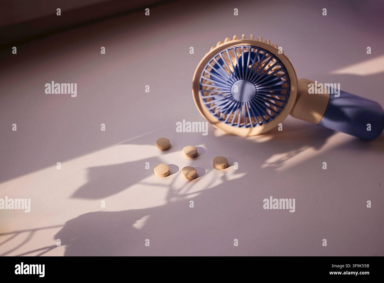 Handheld fan in blue and beige color next to round tablets on a light surface, with soft shadows created by natural light Stock Photo