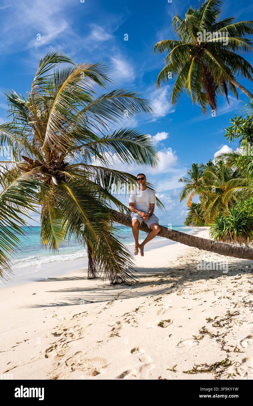 A man sits on a leaning palm tree along a tropical beach in Maldives Stock Photo