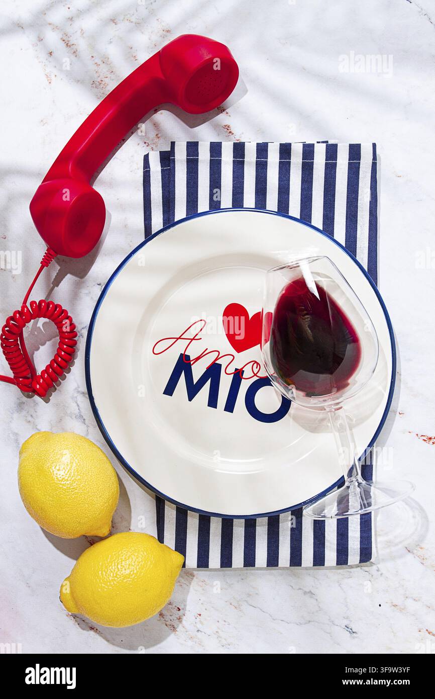 A round white plate sits on a striped napkin in a kitchen. It has the words Amore Mio written on it. A red phone and two lemons are placed nearby Stock Photo