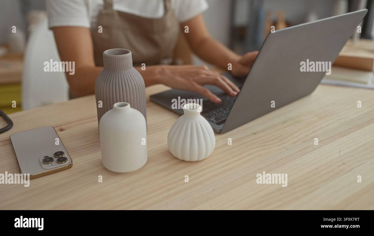 Woman typing on laptop showing hands beside three ceramic vases on wooden table in studio; focus craft calm. Stock Photo