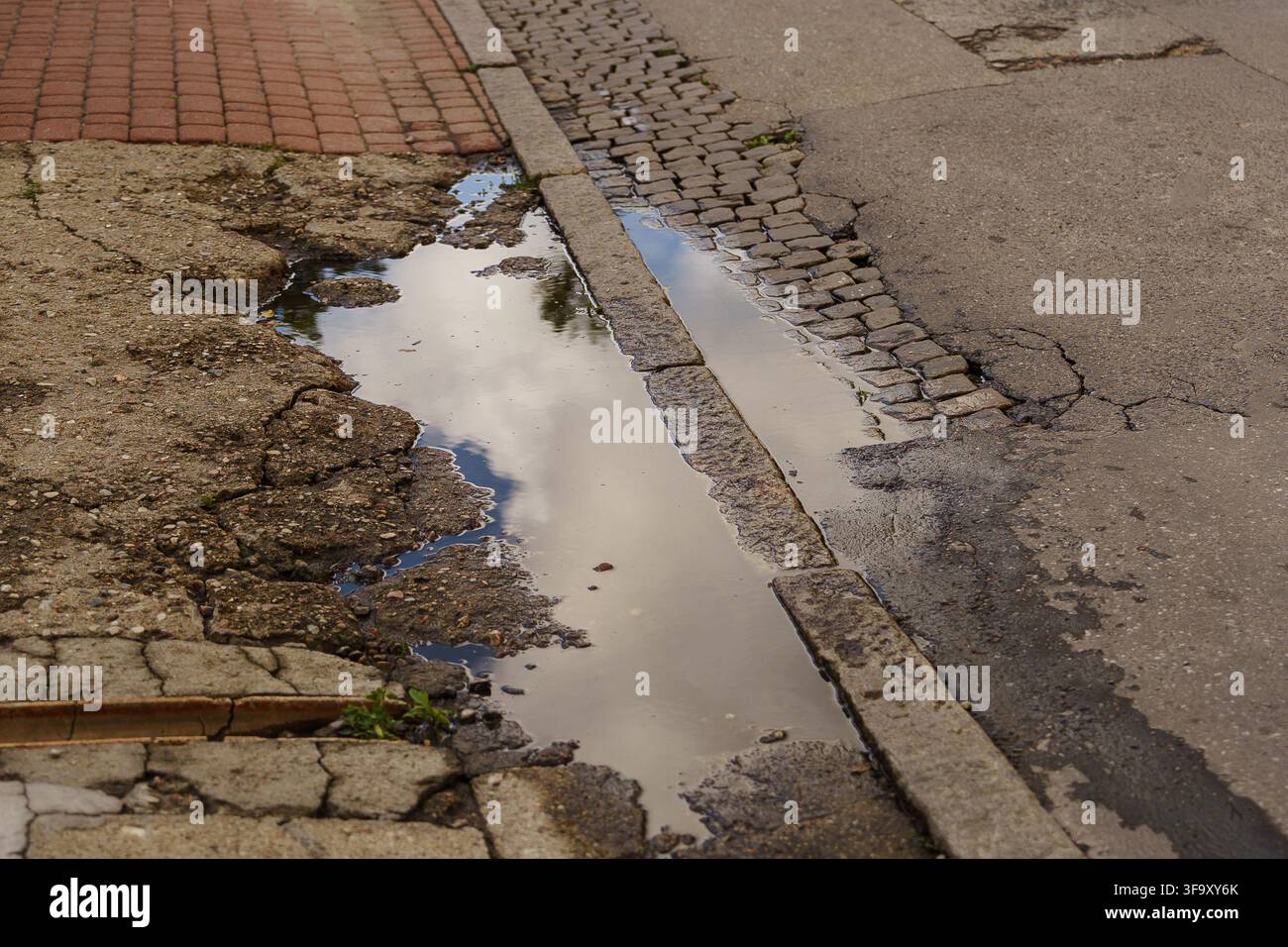 Rain puddle on a cracked, weathered asphalt street, reflecting a cloudy sky, illustrating urban infrastructure decay and road surface texture Stock Photo