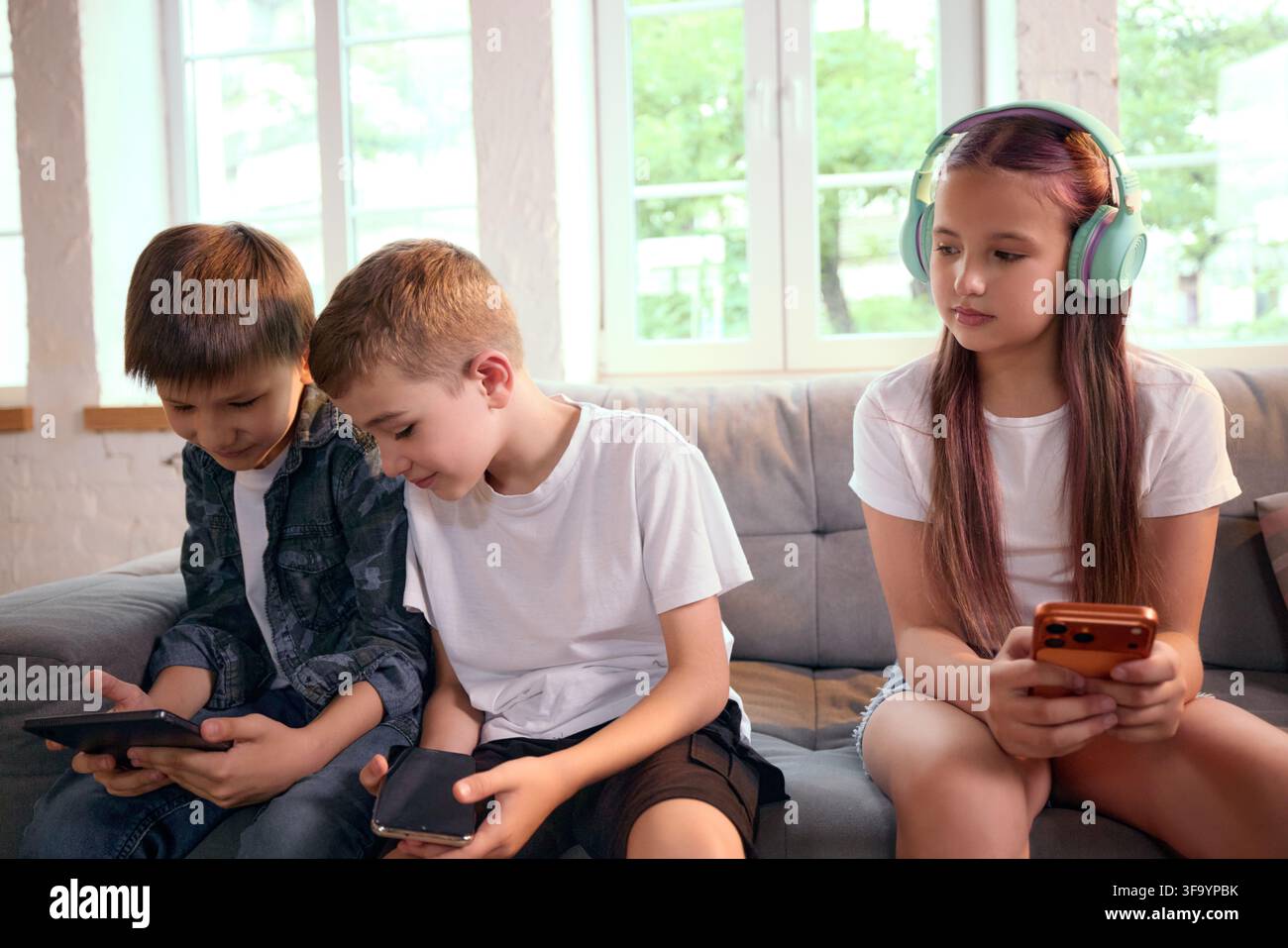 Three school-age friends using personal mobile devices together on sofa Stock Photo