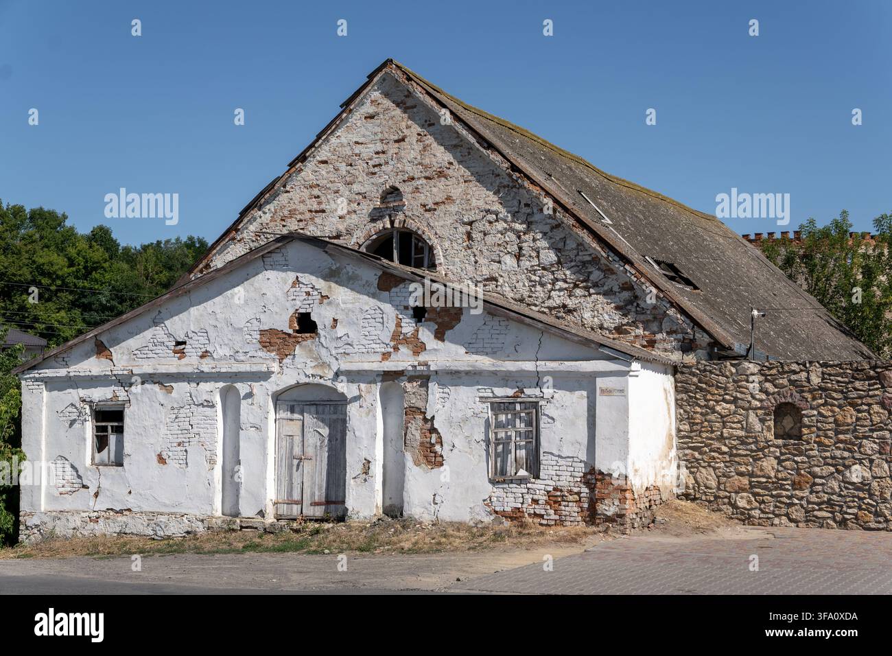 Ancient historical building features peeling white plaster, exposed brickwork, and closed wooden door. Weathered masonry structure stands under clear Stock Photo