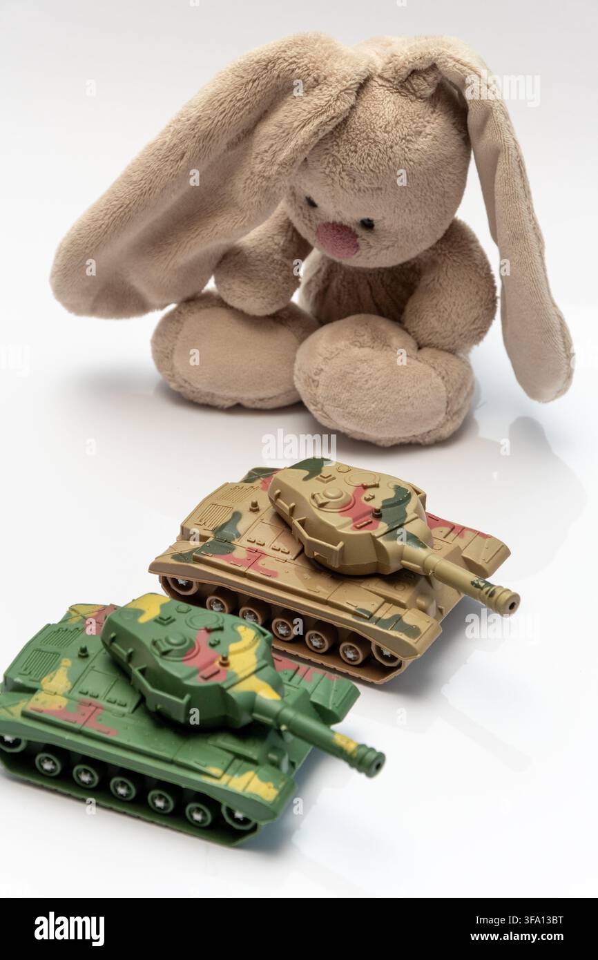 A plush bunny sits behind two camouflage toy tanks on a white background. Stock Photo