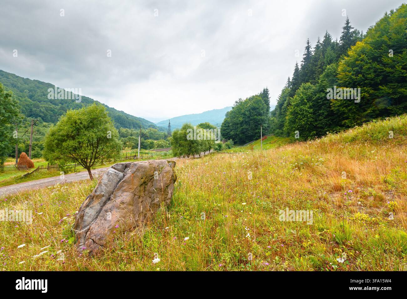 rural landscape. fence near road through field and forest to the high mountains. countryside scenery of ukraine in summer with overcast sky Stock Photo