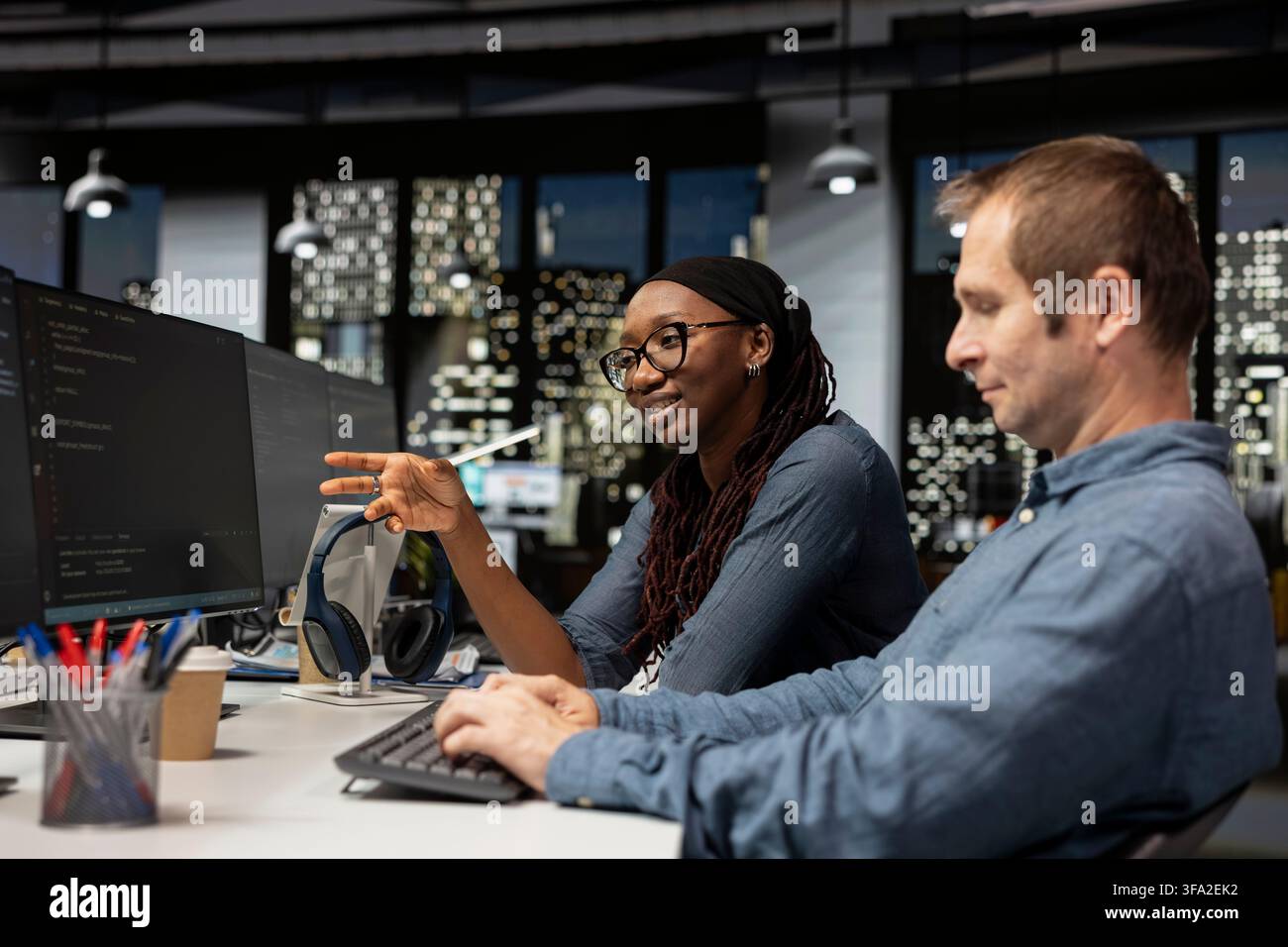 Cheerful tech startup intern asks questions to senior programmer coding advanced AI algorithms and machine learning models enhancing automation. Smiling worker requests help from manager in office Stock Photo