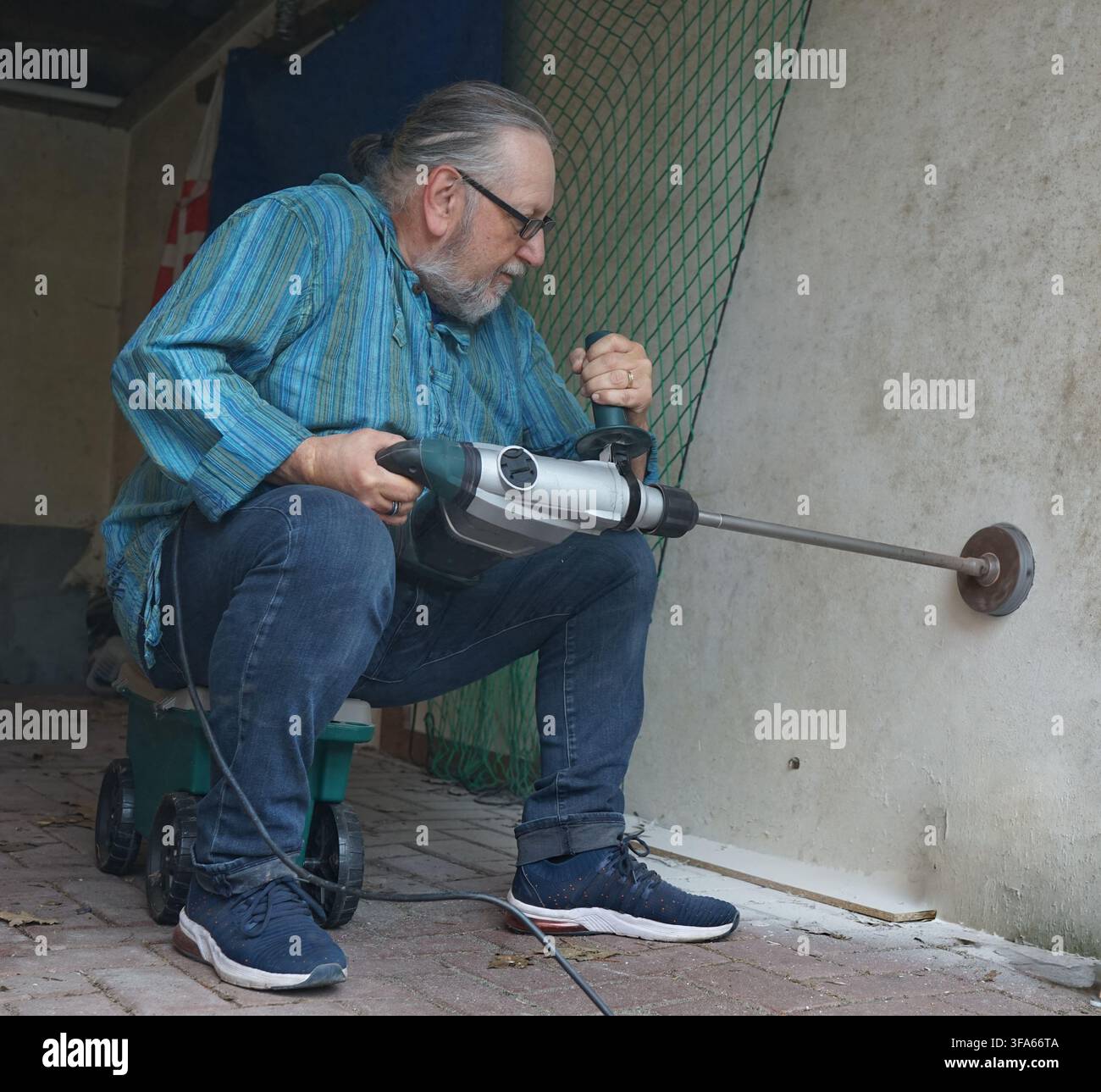 An older man, wearing glasses and a blue striped shirt, is sitting on a wheeled cart while using a jackhammer to bore into a brick wall. He is positio Stock Photo