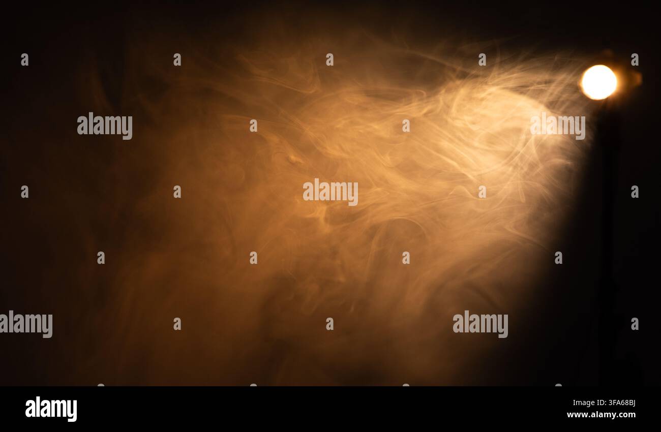 Dramatic warm spotlight beam shining through dense smoke on black background  Stock Photo