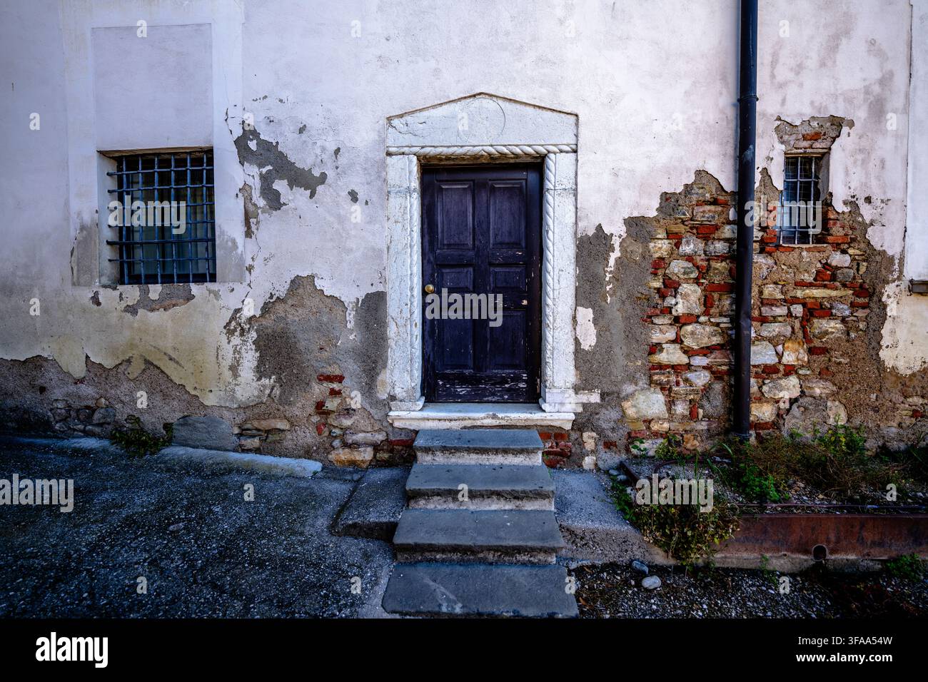 Dark wooden door framed by carved stone in a weathered plaster facade with barred windows, exposed brickwork and worn masonry surfaces. Stock Photo