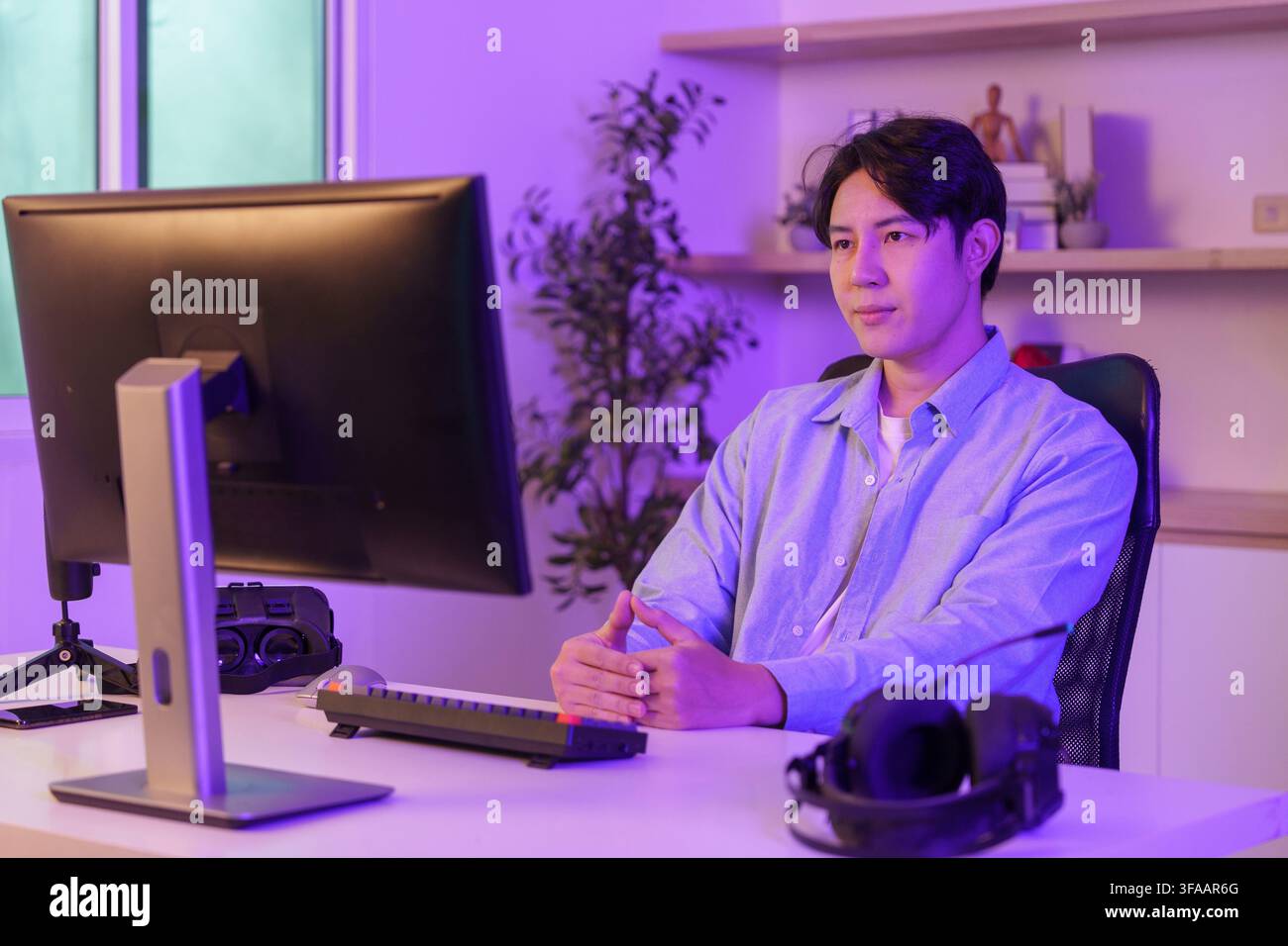 Asian man sitting at desk looking at computer monitor in neon purple lighted home office for remote work Stock Photo