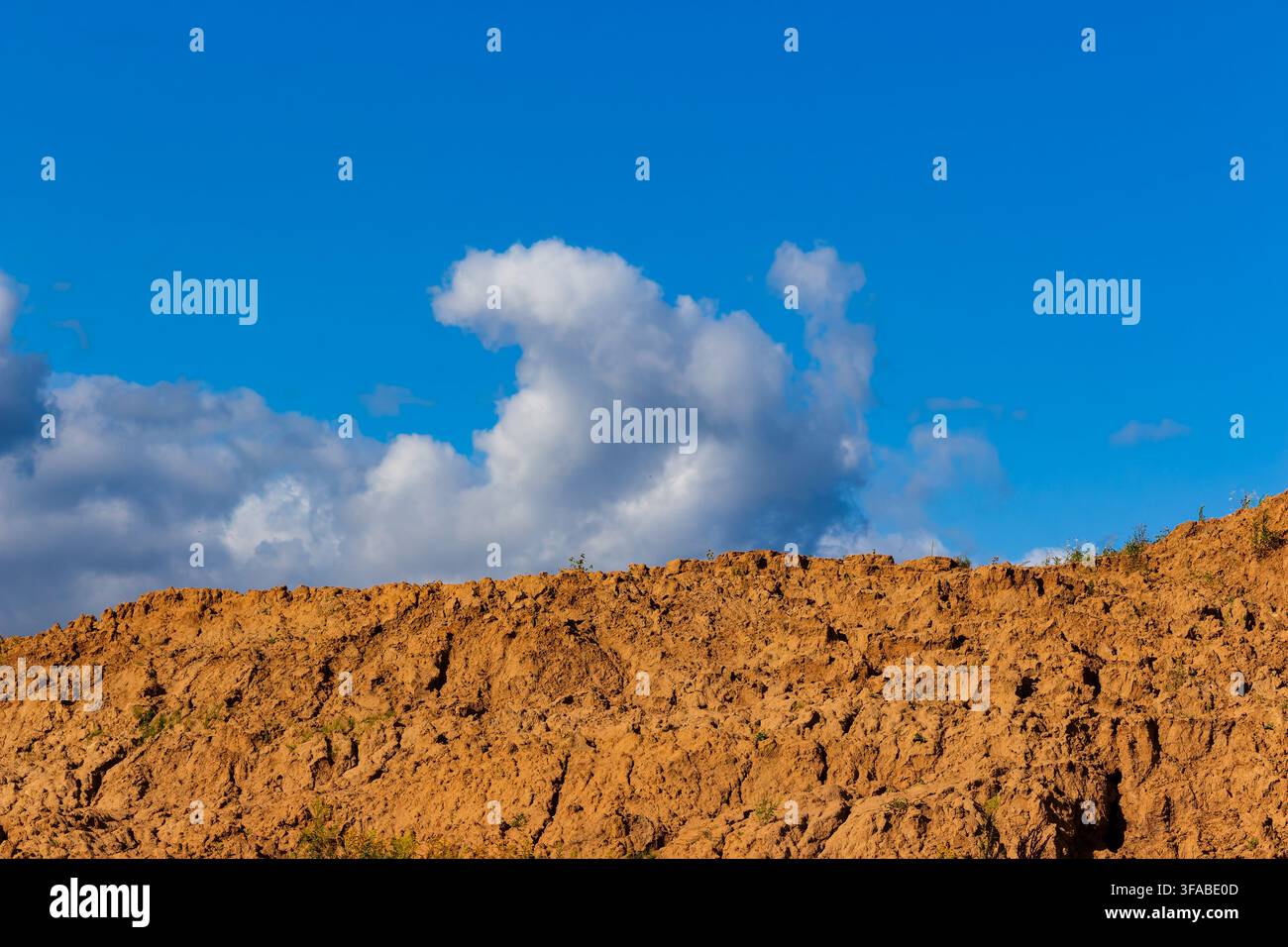 Large piles of excavated brown soil and raw earth mounds set against a vibrant blue sky with fluffy white clouds, showcasing raw land development and Stock Photo