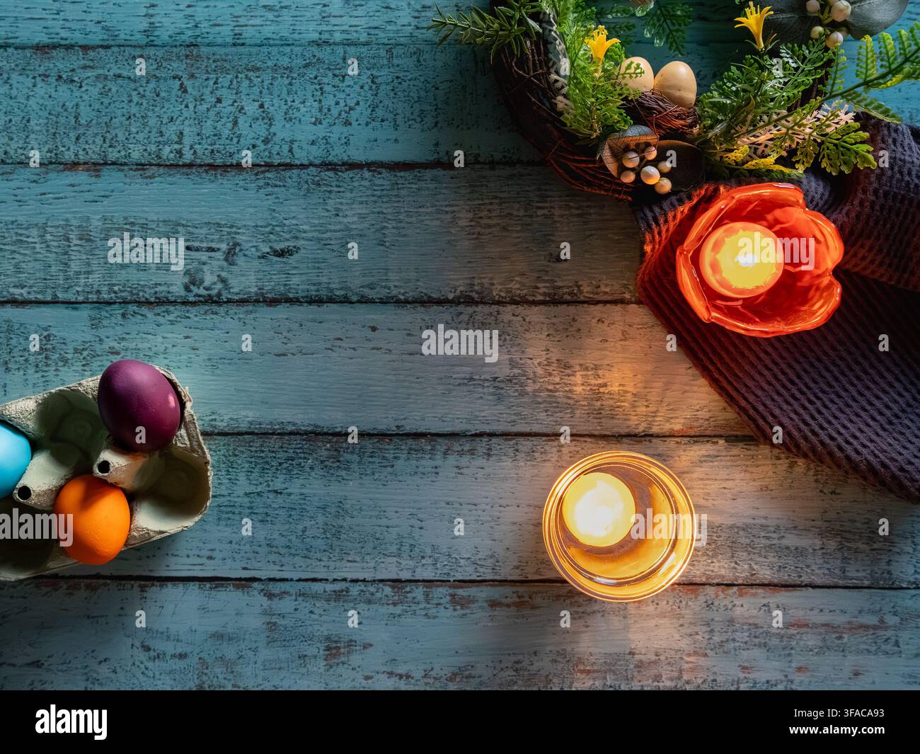 Festive Easter still life featuring a glowing candle decorative eggs spring greenery and a bright orange flower arranged on a blue wooden background. Stock Photo
