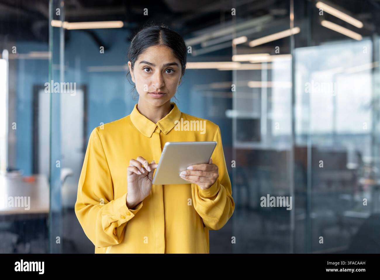Portrait of a young serious Indian woman standing in an office, holding a tablet and looking confidently at the camera. Stock Photo