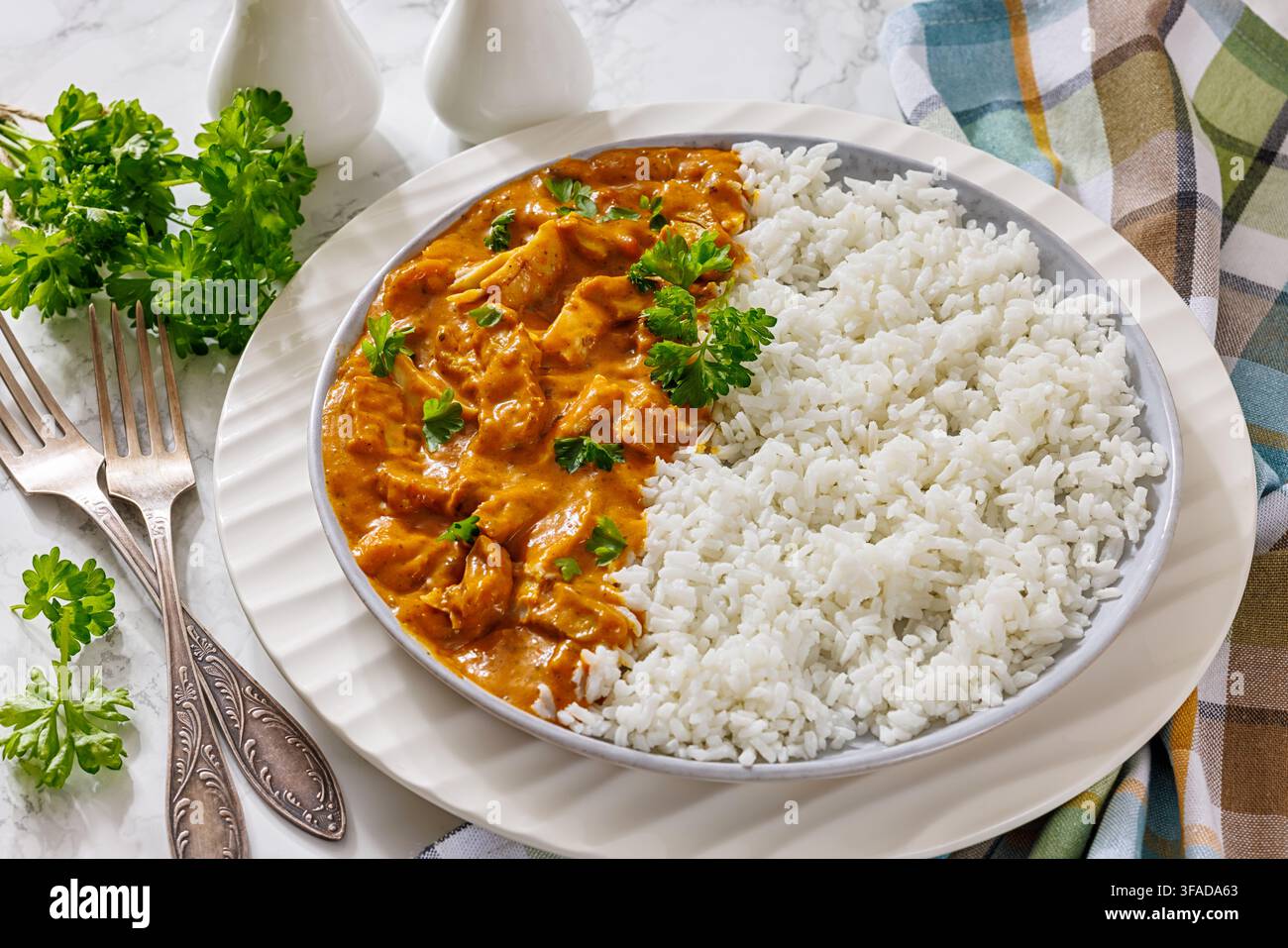 Spiced chicken fillet curry with cooked long grain rice, served on a plate with forks, yougurt, napkin on light marble backdrop, horizontal view, not Stock Photo