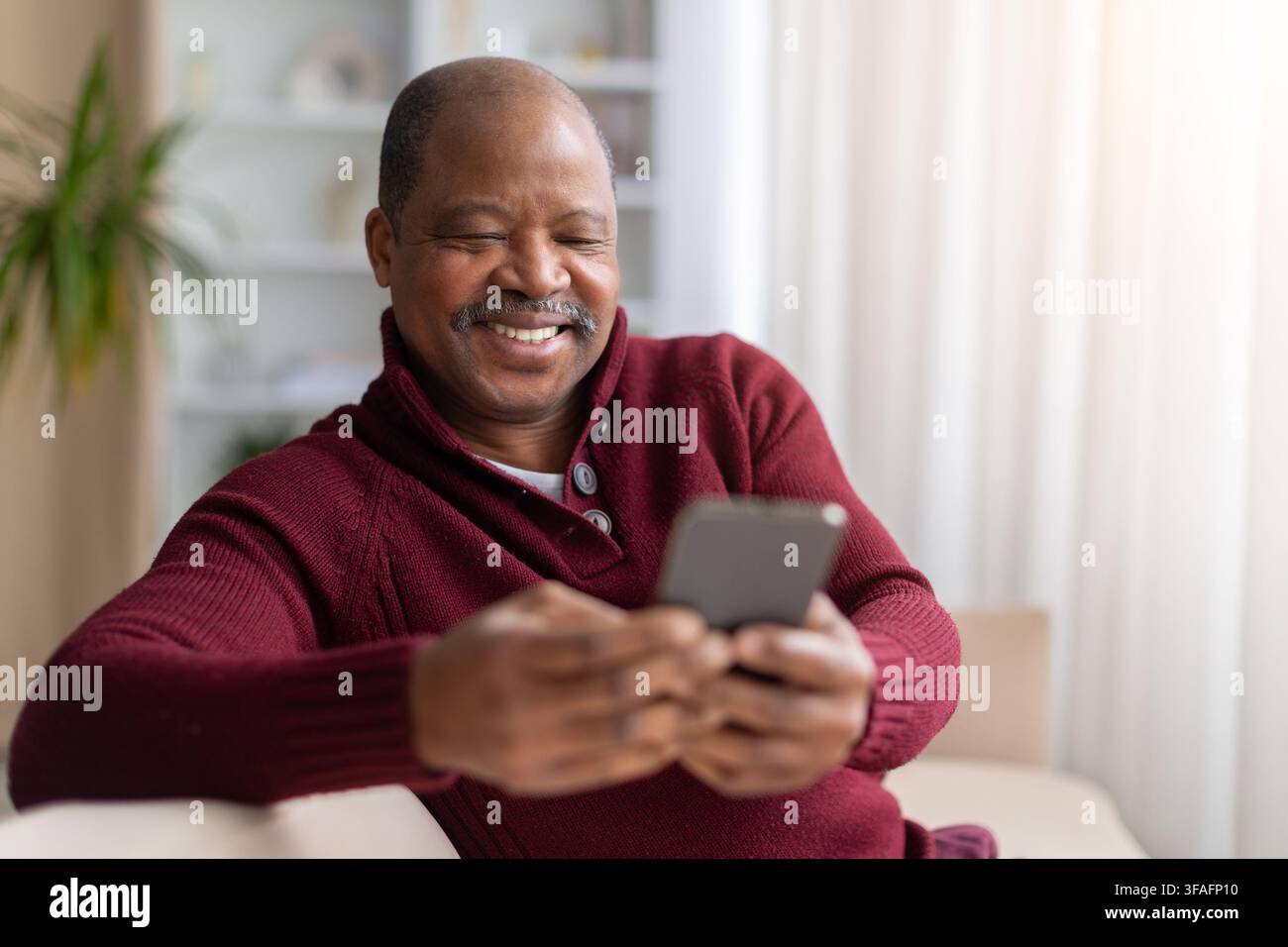 Smiling mature Black man relaxing on sofa at home while using smartphone Stock Photo