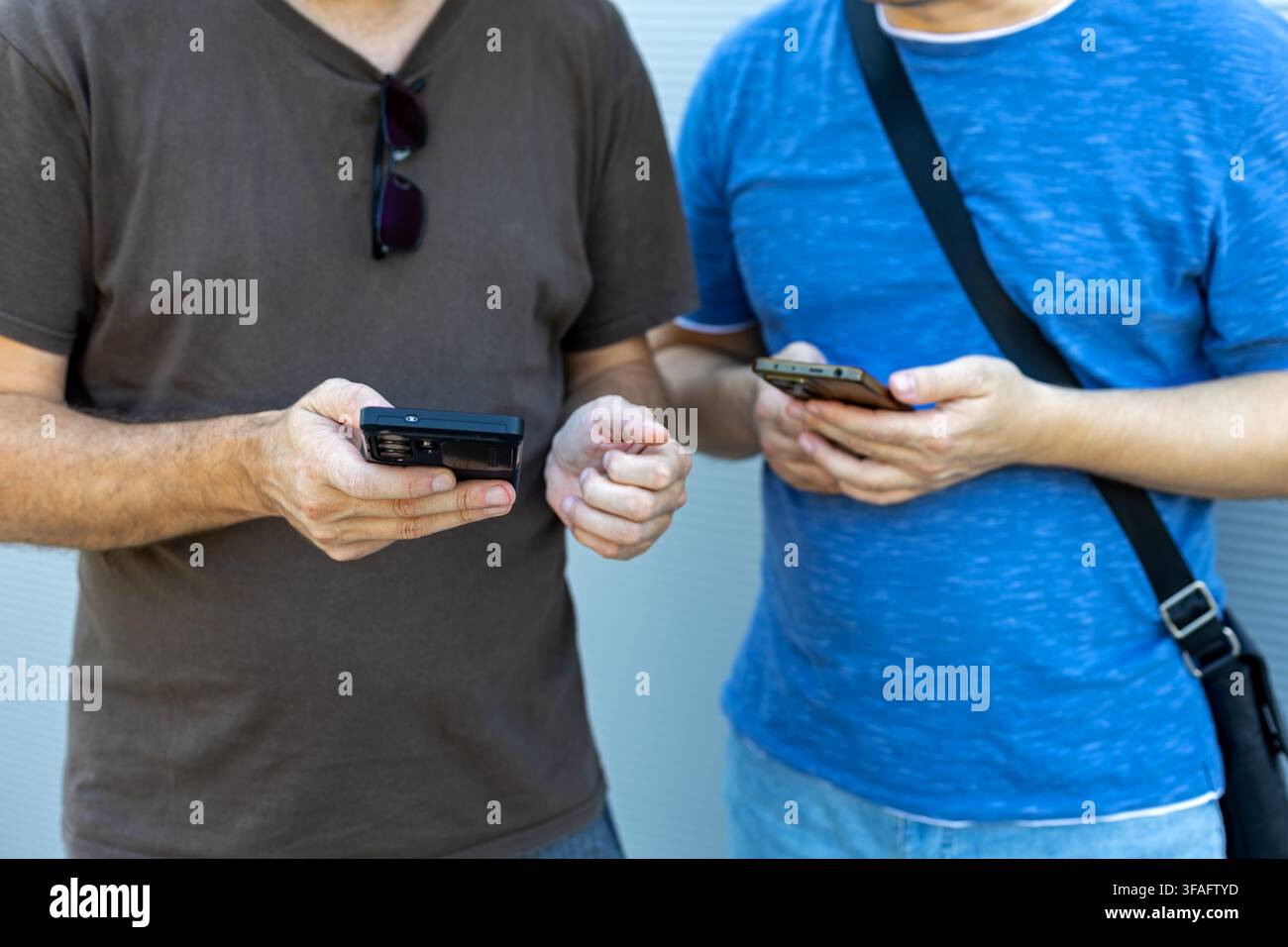Two Men Using Smartphones Side by Side. Two men standing side by side using smartphones while dressed casually against a light neutral background. Stock Photo