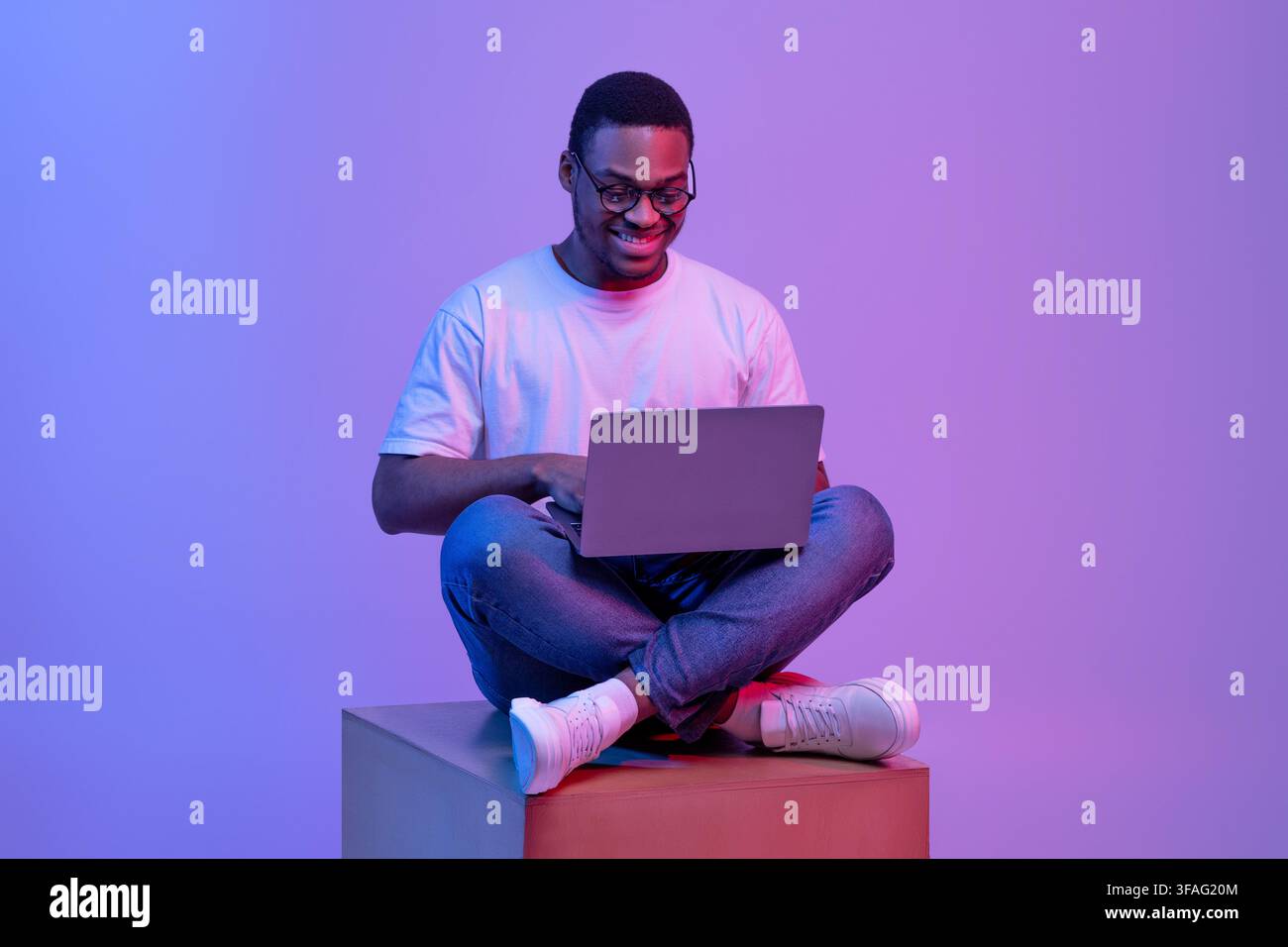 Black Programmer Guy Using Laptop Computer, Sitting On Cube In Neon Lighting Stock Photo