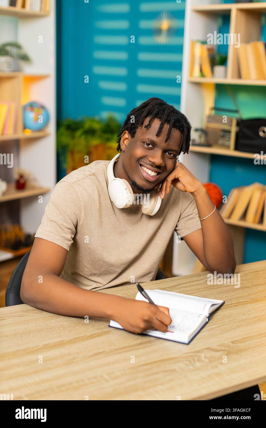 African American young man on home table writes notes in notebook with pen for study planning at home. Black guy on home table reviews written task list with happy focused mood in living room interior Stock Photo