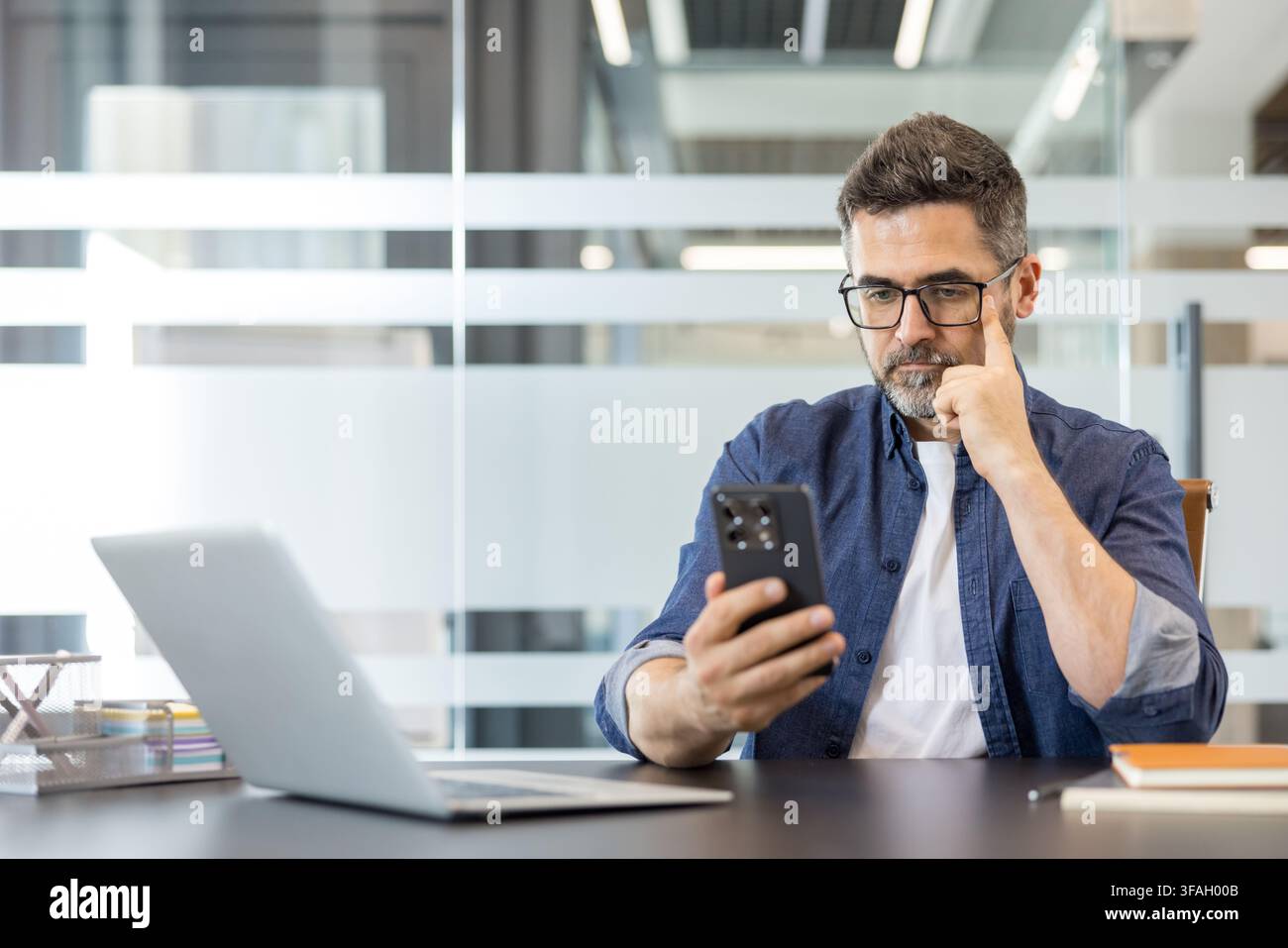 Confident businessman wearing glasses using his mobile phone and laptop at a desk in a modern office, concentrating on digital communication and online tasks Stock Photo