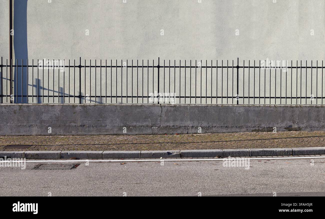 Tubular metal fence on a concrete base at the roadside. Light green plaster wall on behind, sidewalk and street in front. Background for copy space. Stock Photo