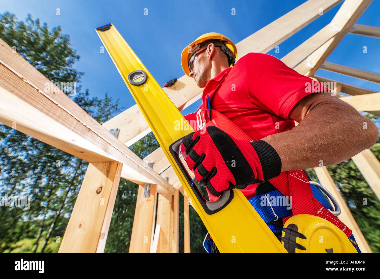 A worker holds a level tool while measuring wooden beams at a construction site. The location has bright skies and trees in the background. The worker Stock Photo