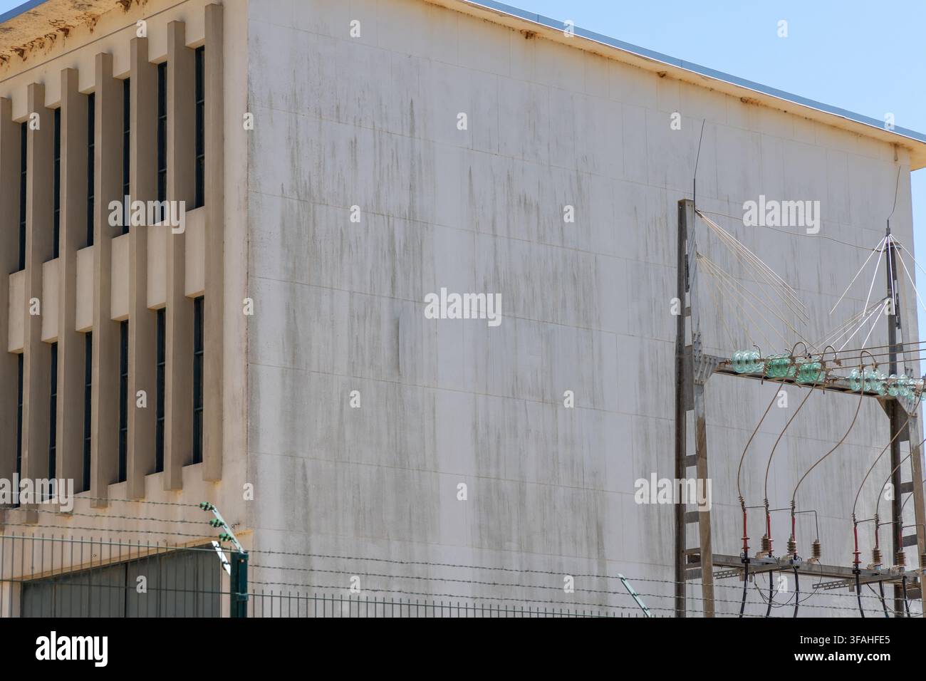 A stark concrete building with a repeating pattern of narrow windows stands beside a weathered utility pole carrying electrical insulators and wires Stock Photo