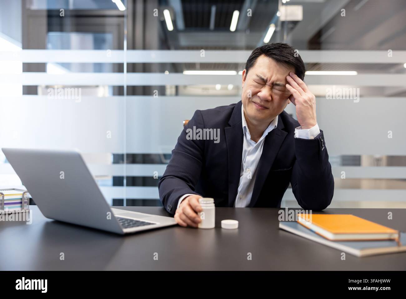 Asian businessman suffering from a severe headache or migraine, feeling stressed and exhausted while trying to work on his laptop in a modern office, holding a bottle of pills Stock Photo