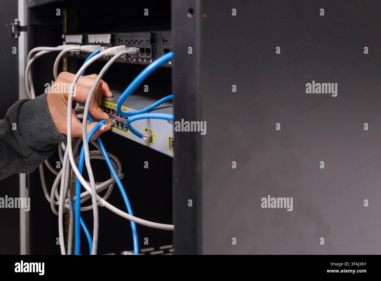 Medium shot view of a technician plugging a blue ethernet cable into a network switch inside a server rack. Stock Photo