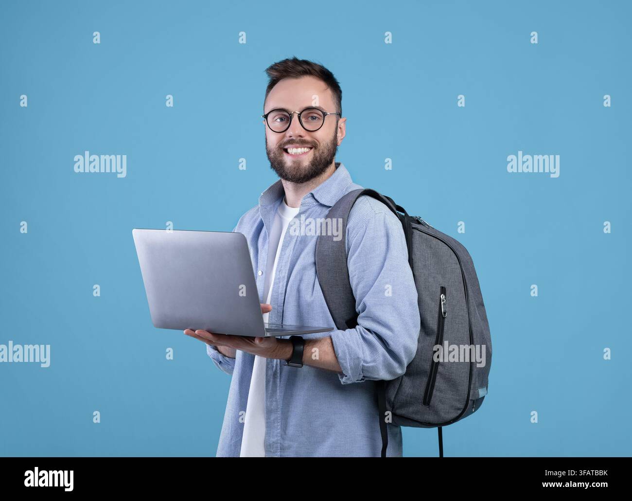 Smart millennial student with backpack using laptop computer over blue studio background Stock Photo
