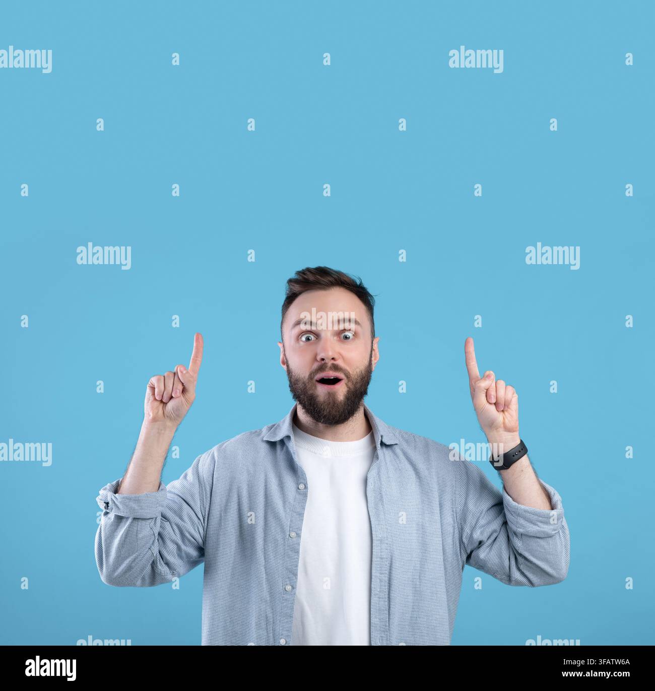 Handsome bearded guy having shocked face expression, pointing at blank space over his head on blue studio background Stock Photo