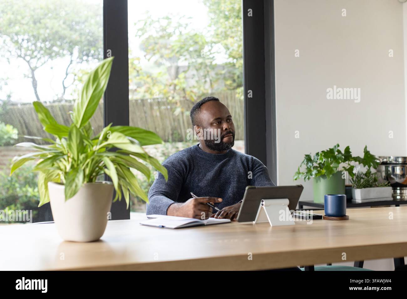 African man sitting at wooden table in home, writing in notebook and using tablet Stock Photo