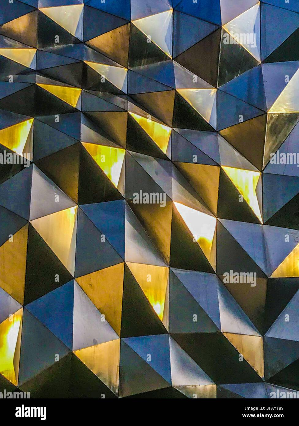 Modern geometric architectural facade with 3D metallic triangular panels reflecting golden light. Stock Photo