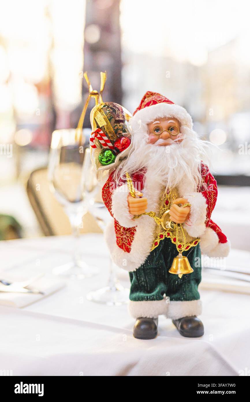 A decorative Santa Claus figurine stands on a table. The Santa has a white beard, red suit, and holds bells and a sack. The background is softly blurr Stock Photo