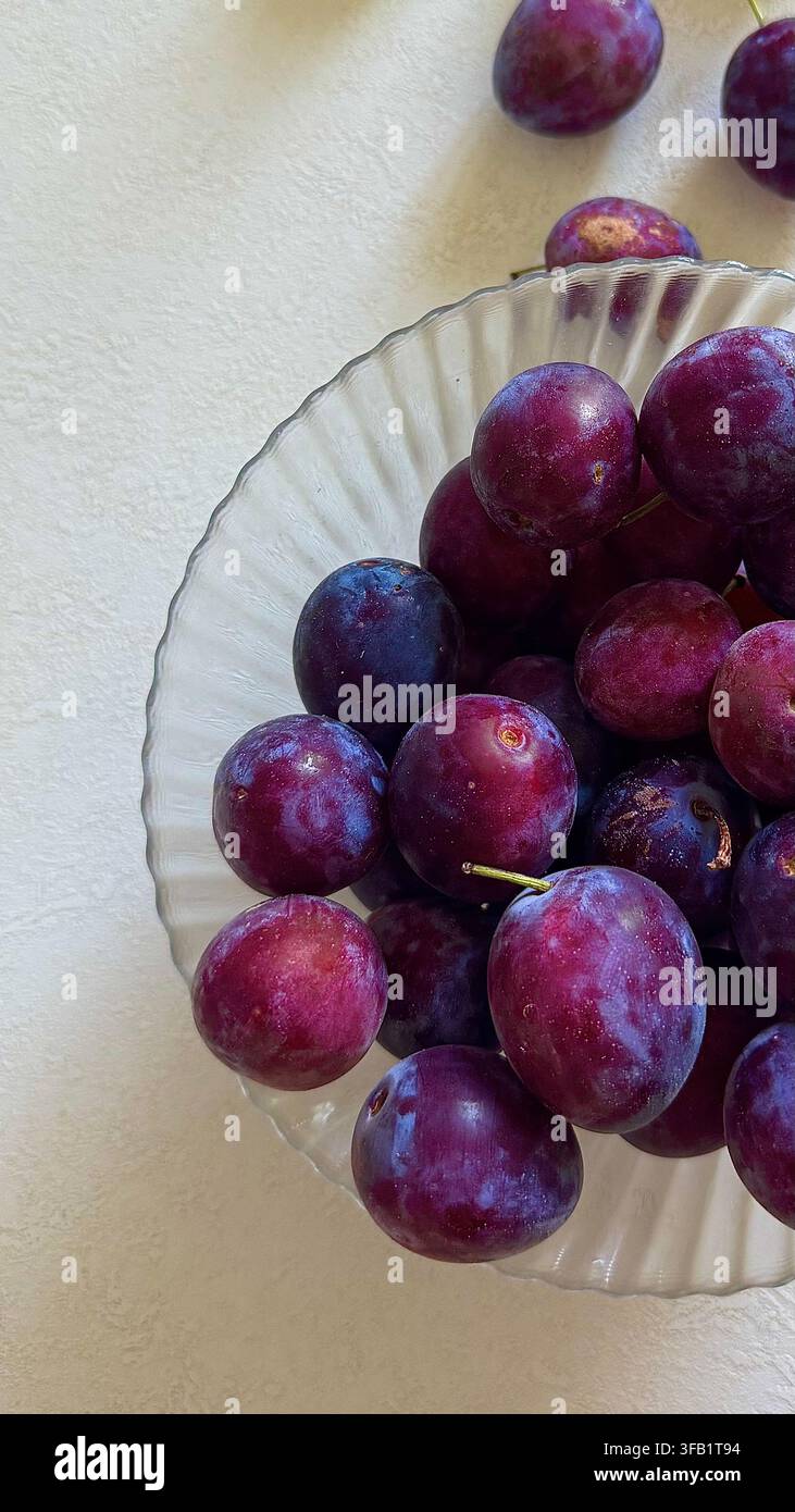 Ripe purple plums arranged in a clear glass bowl, creating a simple fresh fruit still life on a light surface. Stock Photo