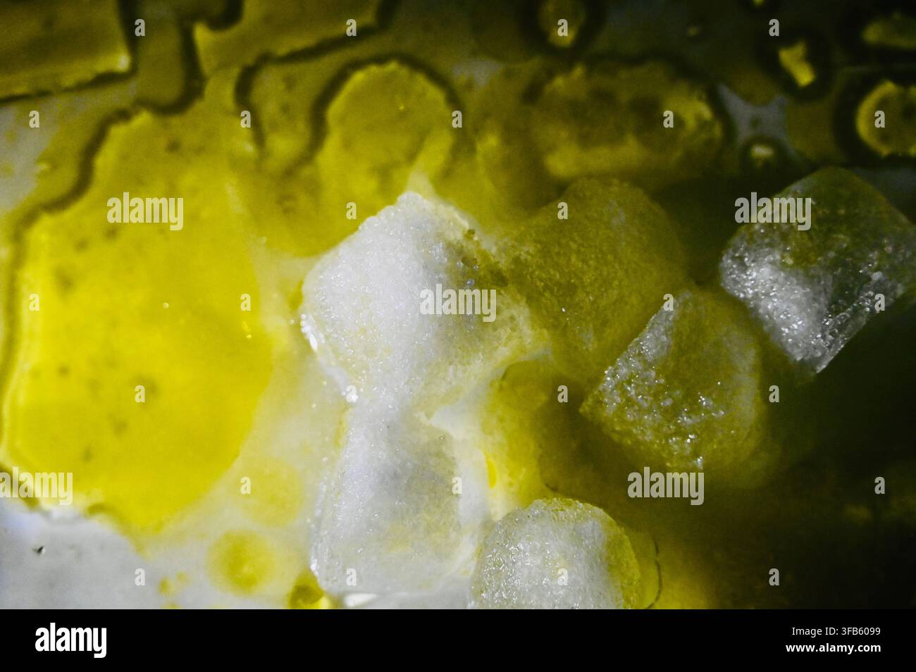 A horizontal studio macro photograph of melting ice cubes surrounded by pools and glistening drops of extra virgin olive oil on a white surface. Stock Photo