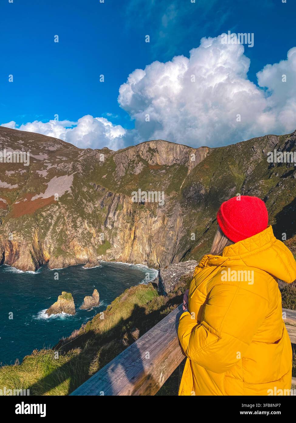 A woman in a red hat and yellow jacket looks out over the sea and theSlieve League/Sliabh Liag cliffs in Donegal, Ireland along the Wild Atlantic Way. - Smartphone Captured Stock Image