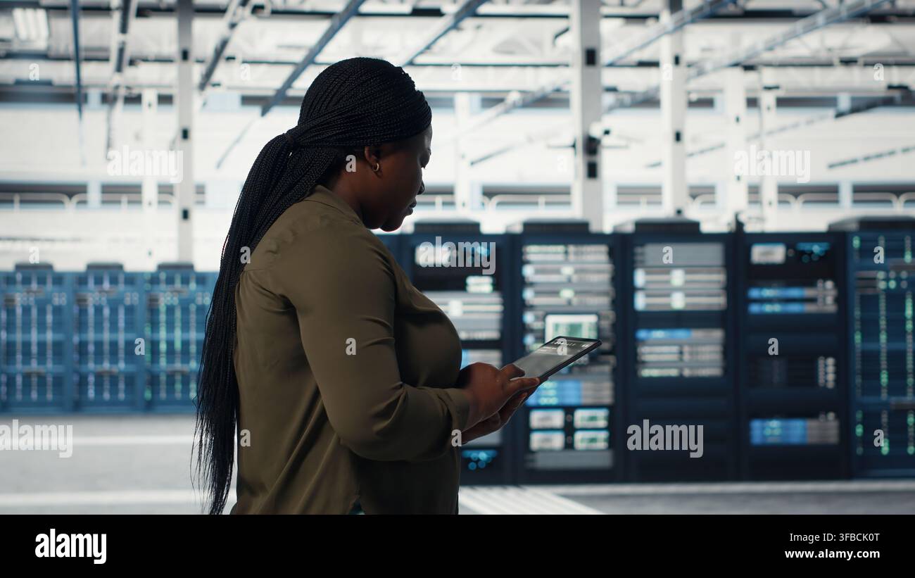 Technician walking in data center with tablet, checking hardware for system stability. Worker overseeing server farm with device, configuring infrastructure rigs, camera B Stock Photo