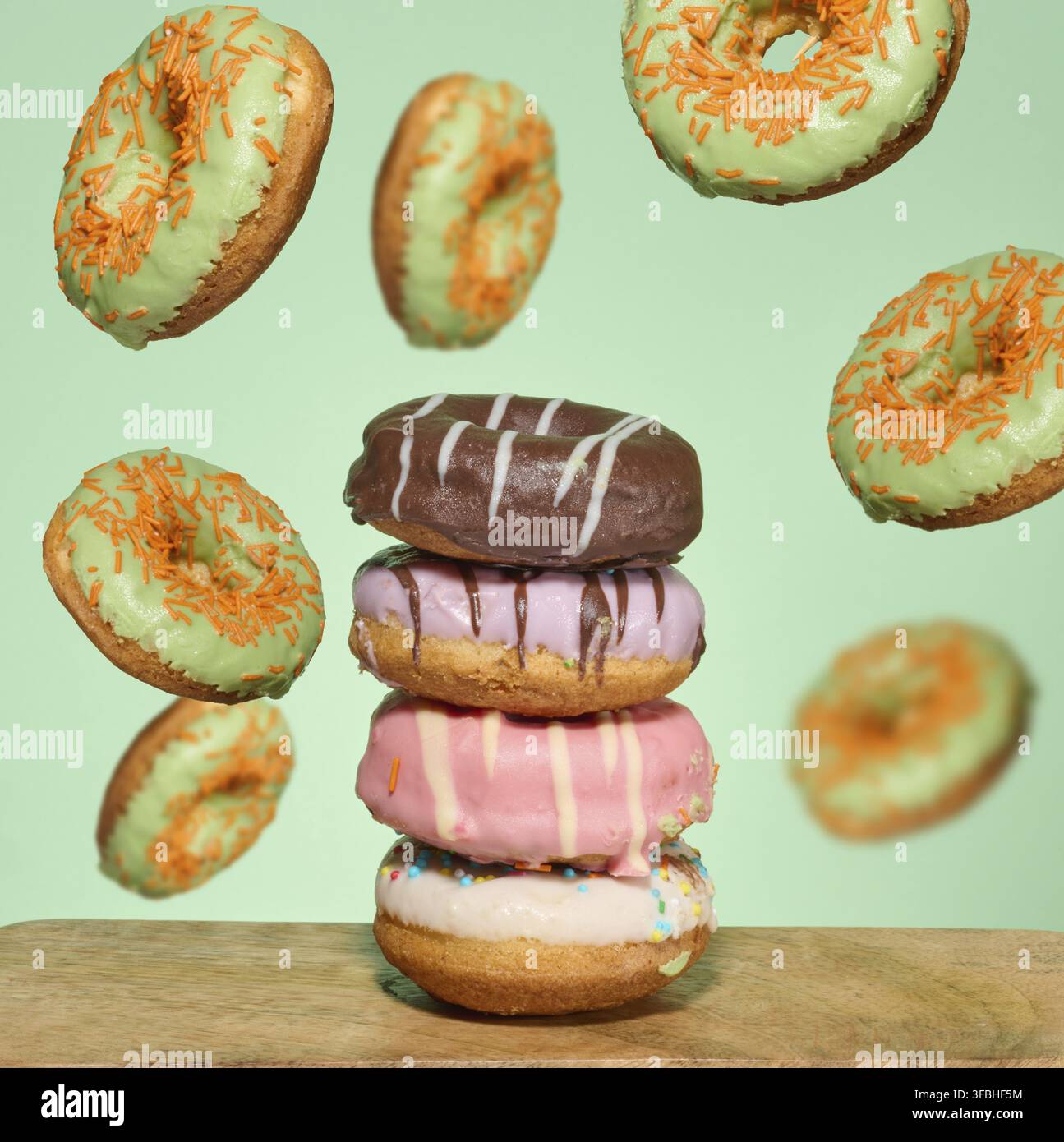 A stack of four brightly glazed donuts on a table with green donuts with orange sprinkles flying around them Stock Photo