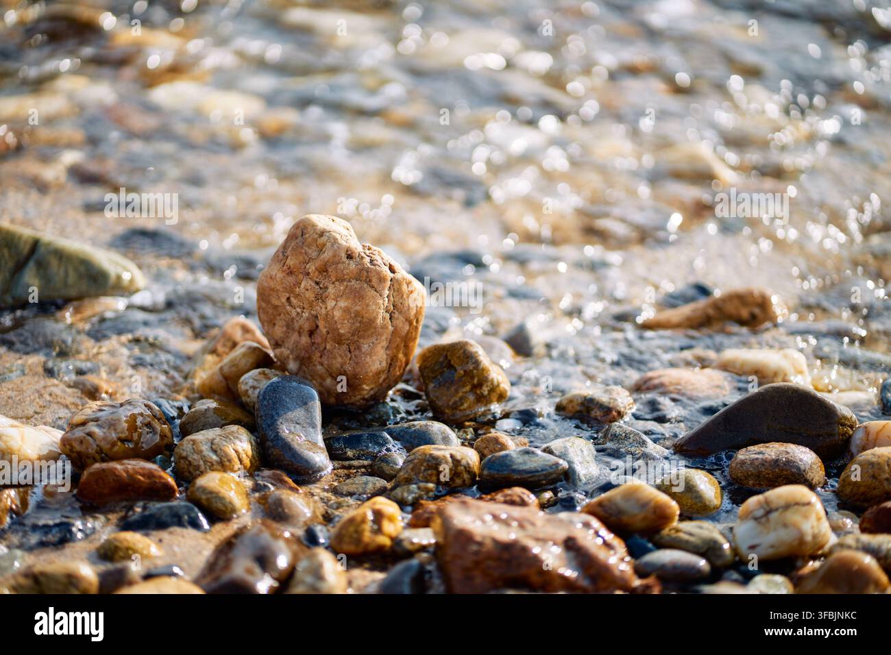 Close-up view of smooth multi-colored pebbles and wet sand on a sea shore with sparkling water ripples washing over the ground in Greece. Stock Photo