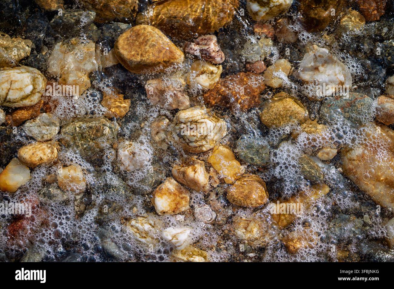 High angle close-up shot capturing wet colorful pebbles and sparkling sea foam washing over a rocky beach shore. Stock Photo