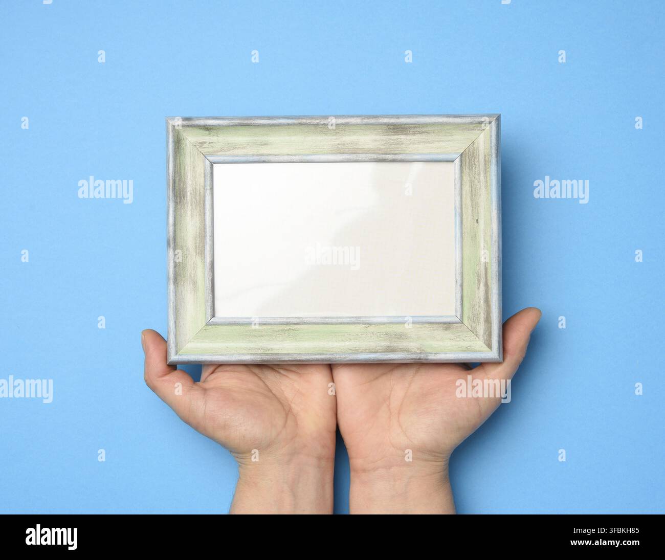 Two female hands holding a rectangular blank wooden frame, blue background Stock Photo