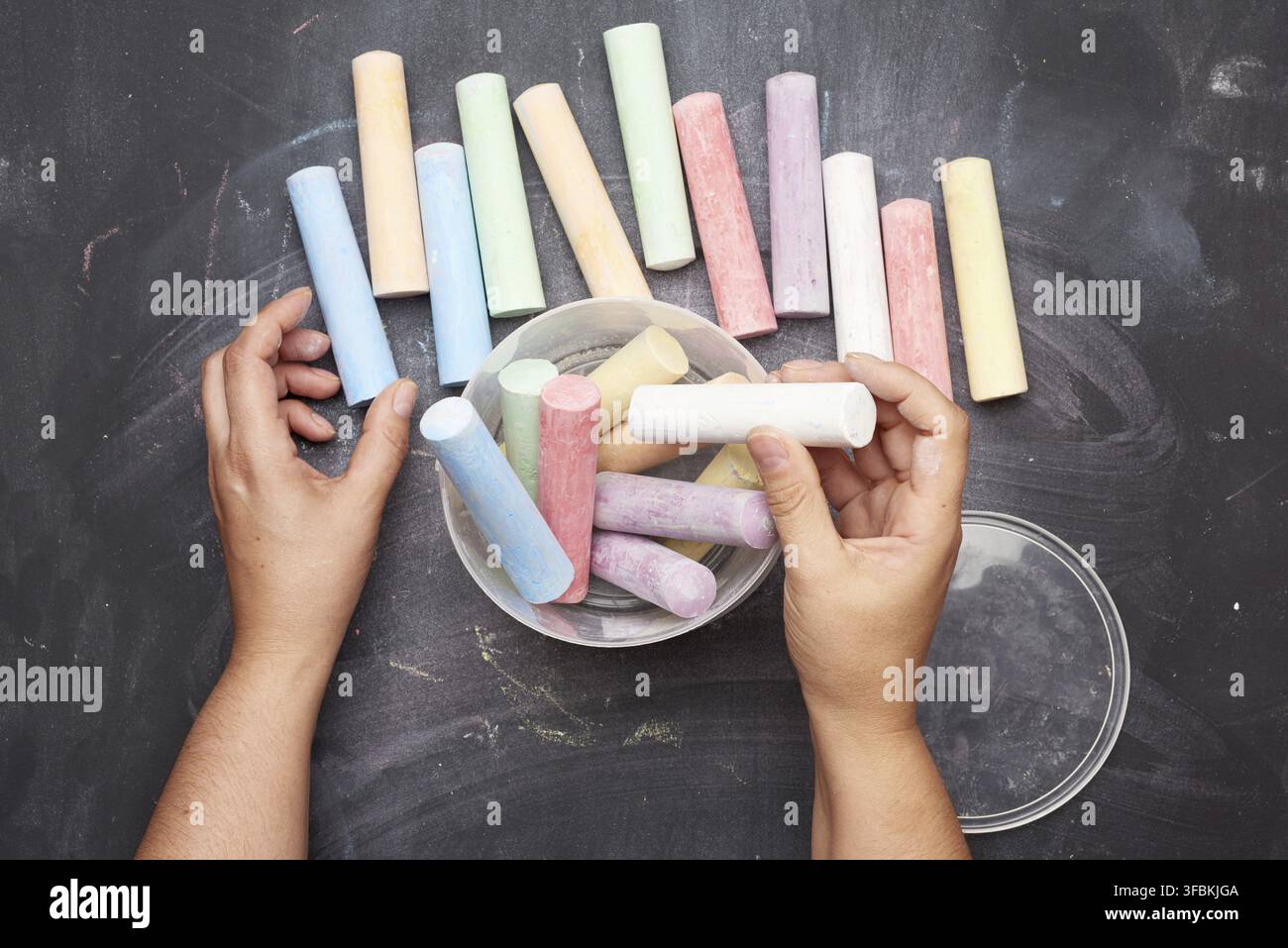 Multicolored chalk in a plastic bucket on a black background, female hands fold objects into a container, top view Stock Photo