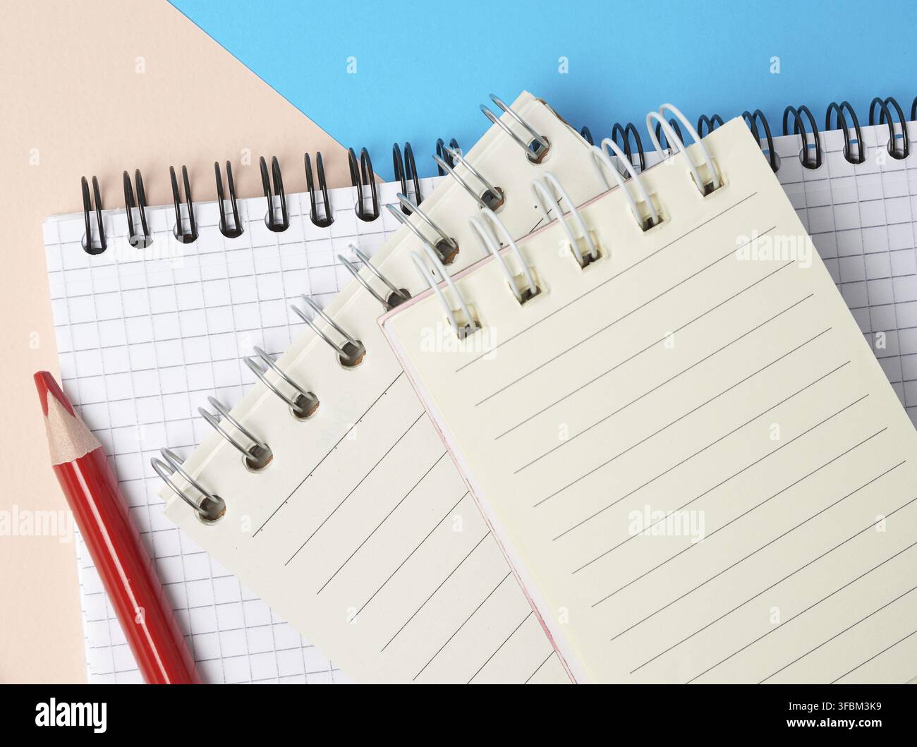 Red wooden pencil and stack of spiral open notebooks with blank white sheets on a blue background, top view Stock Photo