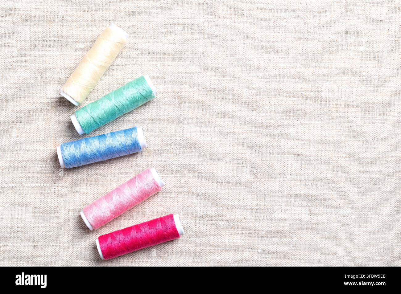 Polyester sewing thread spools randomly distributed on linen fabric, from above, with copy space. Multi colored vibrant polyester sewing thread. Stock Photo