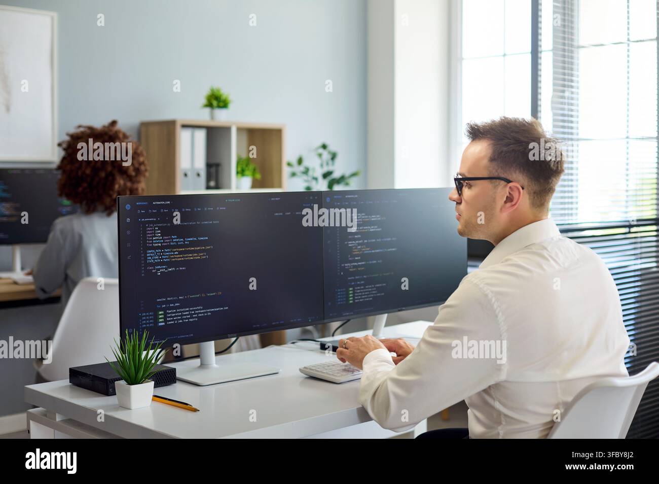 Software developer coding at dual monitors in modern office workspace, daylight Stock Photo