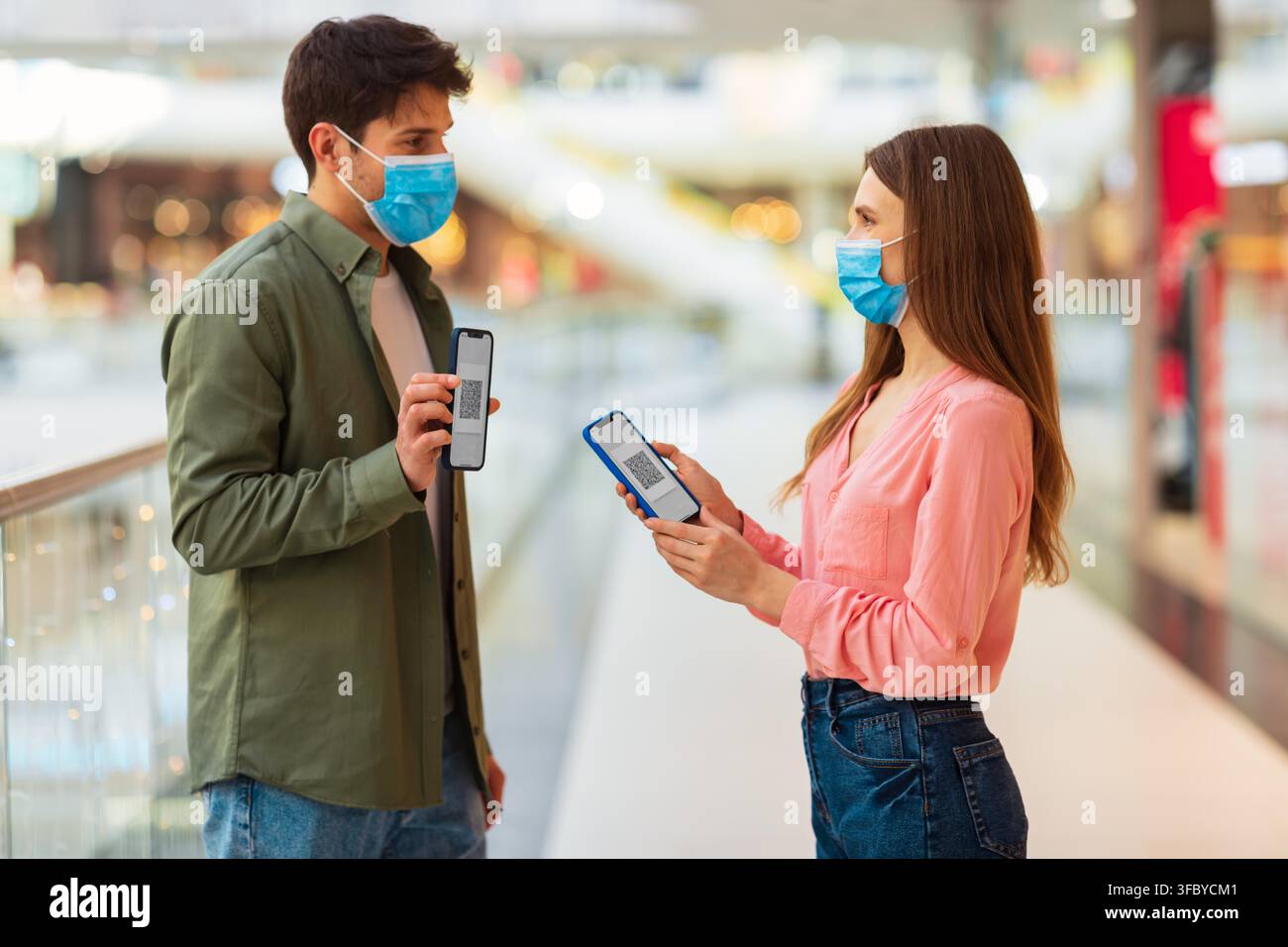 Happy Young Couple Using Smartphones In Shopping Mall, Wearing Face Masks, Shopping Concept Stock Photo
