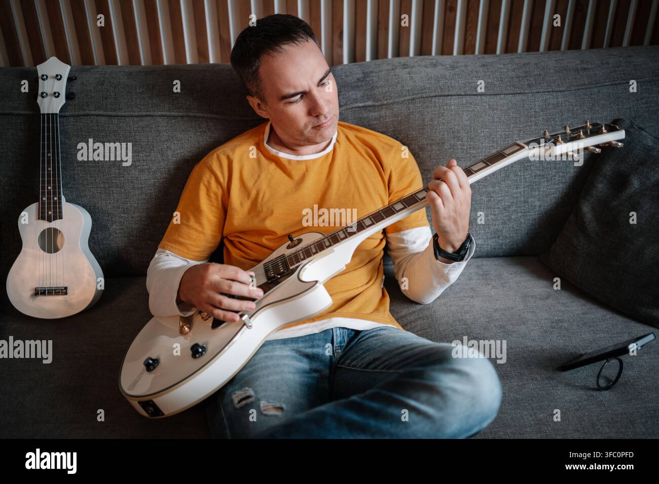 Man playing guitar while sitting on sofa, musical hobby and relaxed home atmosphere. Stock Photo