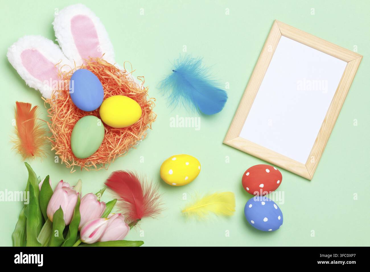 Easter eggs in a nest, bunny ears, flowers and a frame with copy space on a mint green background Stock Photo